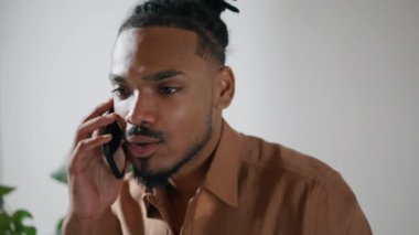 Aydınlık odada akıllı telefondan konuşan dikkatli bir adam. Çağdaş serbest çalışan cep telefonuyla iş haberlerini tartışıyor. Odaklanmış adam ofisten cep telefonunu arıyor. Afro insan olumlu konuşuyor. 