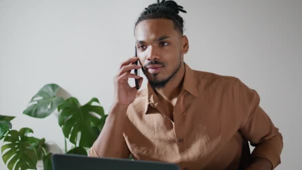 オフィスの近くで携帯電話をチャット忙しい男 アフリカ系アメリカ人のフリーランスは 自宅の職場でノートパソコンの通話を終了 黒髪の男がリモートで働いて電話を拾う 集中型ヒップスター型キーボード — ストック動画
