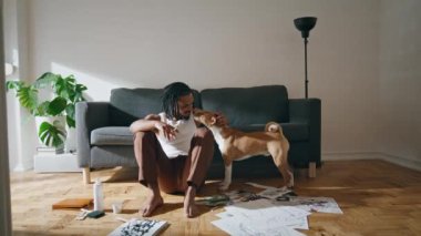 Kanepe odasında pozitif sanatçı köpeği okşuyor. Afrika kökenli Amerikalı bir adam ahşap zeminde oturan evcil hayvanı okşuyor. Yaratıcı adam evde çizimler yaptıktan sonra rahatlıyor. Ressam hobisiyle dinleniyor.