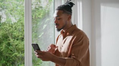 Pencerenin yakınındaki ciddi bir sanal arama mobili. Afrikalı Amerikalı adam videolu sohbet kullanarak açıklıyor. Evde cep telefonuyla konuşan yakışıklı bir adam. Akıllı öğrenci online akıllı telefon konferansı 