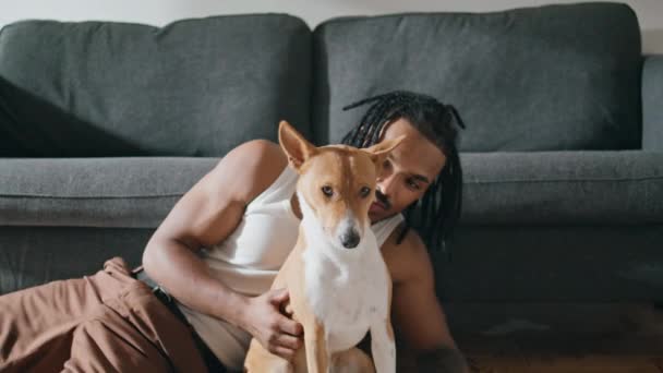 快乐的男人在家里的特写镜头里摸狗 在时髦的公寓里拥抱家养动物的胆小的家伙 一个人在那里缩成一团 非洲裔美国人的主人在客厅里戏弄他的狗朋友 — 图库视频影像