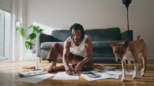 自宅で絵を描くのを楽しんでいる真剣な男 現代人は一人で座って新しいアートワークを作成します 中規模の犬の部屋で歩く アフリカ系アメリカ人のアーティストが趣味に関わる紙でスケッチ — ストック動画