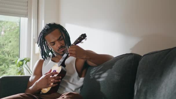国内でギター演奏を中心に活動 現代人はソファに座ってウクレレの文字列に触れる アパートで音楽を楽しむ満足した男 アフリカ系アメリカ人の男家で音を抽出 — ストック動画