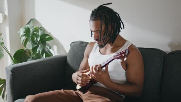 有表现力的吉他手在国内演奏四弦琴 非洲裔美国音乐家坐在沙发上触摸弦乐 黑头发的男人在白色的公寓里放松地欣赏音乐 男人的手抽音 — 图库视频影像