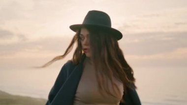 Carefree attractive girl spinning around relaxing on beautiful coast at autumn sunset close up. Relaxed stylish woman in hat posing at nature sundown looking camera. Freedom beauty relaxation concept.
