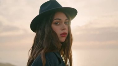 Relaxed girl traveler posing in front beautiful sunset wearing stylish hat close up. Attractive serious woman looking camera confidently enjoying rest on nature. Portrait of pretty model at cloudy sky