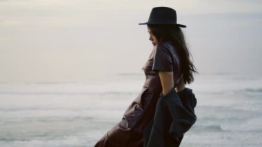 Stylish romantic woman posing at night beach wearing elegant hat. Attractive young brunette sitting chair in front gloomy dramatic seascape. Confident beautiful girl enjoy overcast nature at evening.