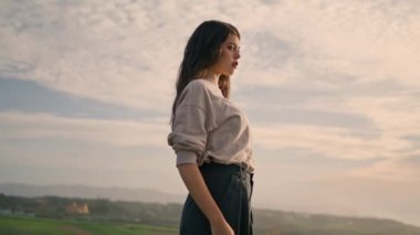 Beautiful young model in front cloudy sky summer evening. Confident attractive woman posing at nature keeping hands in pants pocket. Stylish serious girl standing on valley looking amazing landscape.