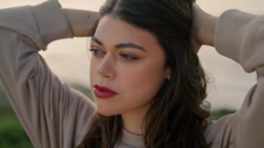 Portrait of confident beautiful model looking camera at seacoast. Gorgeous girl posing on beach hill touching hairstyle. Attractive woman face with seductive red lips magnetic dark eyes close up.