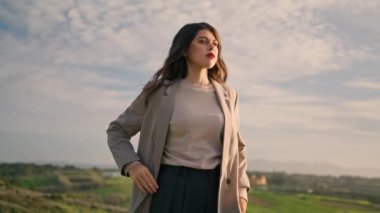 Elegant dark-hair girl posing confidently in front amazing cloudy sky. Attractive young woman in stylish clothes looking camera putting hands at waist. Gorgeous model standing on beautiful green hill.