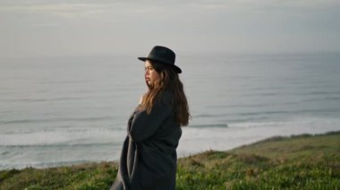 Attractive serious woman standing cloudy seacoast in front gray ocean waves. Young confident girl posing on green grass shore looking camera. Long-hair brunette wearing elegant hat dark coat outdoors.