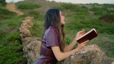 Dreamy romantic girl reading book sitting at nature leaning on rocky hill close up. Attractive young brunette wearing dark dress enjoy hobby outdoors gloomy autumn weather. Relax leisure concept.