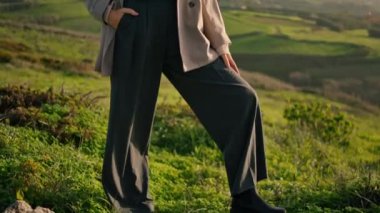 Stylish confident woman standing green grass in front beautiful cloudy sky. Attractive serious girl posing at sunlight wearing comfortable casual clothes on valley. Dark-haired model looking distance 