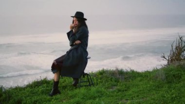 Young fashion woman sitting chair at seashore in front foamy ocean waves wearing stylish hat evening time. Calm model posing on gloomy beach at twilight. Pensive attractive girl taking off dark coat.