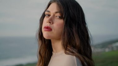 Close up sexy woman face with natural make-up in front gray autumn sky. Portrait of attractive young girl looking camera confidently standing at beach hill. Beautiful model posing on seashore alone.
