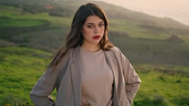 Seductive brunette girl posing on nature looking camera close up. Attractive red lipped woman standing on green grass hill wearing stylish clothes. Portrait of sexy model enjoying beautiful scenery.