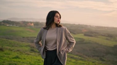 Gorgeous serious woman walking hill wearing stylish clothes close up. Attractive dark-haired girl looking on beautiful scenery with cloudy sky. Confident model posing on nature standing green grass.
