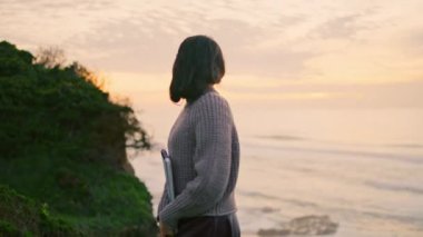 Dreamy pensive woman enjoy ocean twilight walking on coast hill holding album. Attractive thoughtful girl posing in front beautiful sunset sky. Young meditative brunette relax on gloomy nature alone.