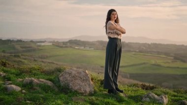 Pensive young woman posing at beautiful valley in front gloomy cloudy sky. Dark-haired attractive girl standing on green grass with hands crossing enjoying amazing landscape. Lady relax on nature.