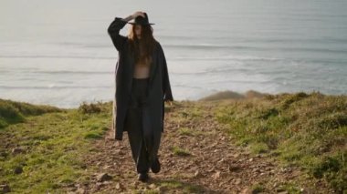 Relaxed stylish girl walking green stony coast at sunny autumn evening. Young woman wearing elegant coat going on grass holding hat from windy weather. Happy brunette enjoying beautiful view ocean