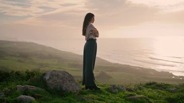 年轻迷人的女子站在青草小山上 望着美丽的落日海景 松驰的长发沉思模型 摆出沿海的姿势 女孩们穿着时髦的休闲装 双手搭在身上 — 图库视频影像