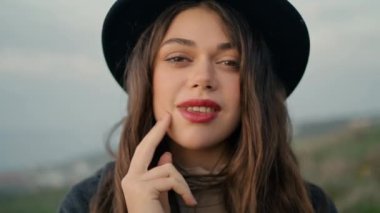 Smiling fashion woman face with red lips at gloomy nature close up. Portrait of confident gorgeous girl wearing stylish hat looking camera outdoors. Happy sensual brunette relaxing enjoy weekend.
