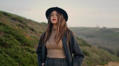 Calm woman traveler looking around hills walking on dirty road alone close up. Attractive young girl in stylish hat spending autumn weekend at countryside. Serious brunette lady enjoy gloomy landscape