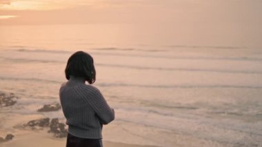 Lonely melancholy girl looking on cold gray ocean waves standing alone at seashore autumn evening. Sad attractive brunette girl posing on empty beach wearing sweater. Loneliness tranquility concept.