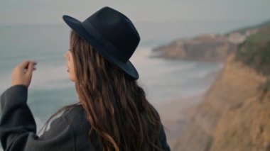 Pensive relaxed girl traveler enjoy seascape standing alone at seashore close up. Attractive brunette turning face to camera touching elegant hat. Young beautiful model posing on nature cloudy day.