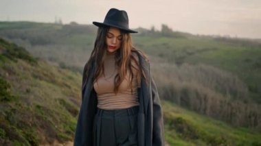 Lonely beautiful woman walking rural road in elegant hat closeup. Attractive fashion girl spending vacation in countryside enjoying autumn landscape alone. Pretty model looking camera. Travel concept