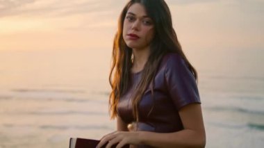 Tranquil charming girl reading book at beautiful sunset close up. Attractive brunette holding foliant standing at seashore alone autumn evening. Young relaxed woman enjoy calm leisure on nature.