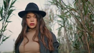 Serious confident woman posing at field with high dry grass cloudy day close up. Attractive bored brunette looking camera wearing elegant hat dark coat. Long-haired tranquil girl standing at nature.