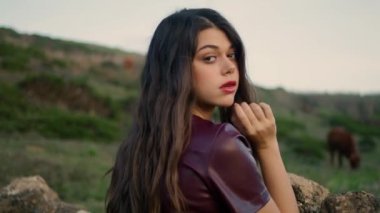 Young woman elegant standing pasture looking on cute brown cow gazering eating green grass. Attractive brunette posing standing near stone fence at gloomy nature close up. Rest relax leisure concept.