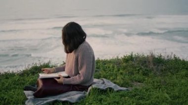 Lonely brunette sitting on blanket with book looking gloomy seascape alone. Attractive young woman relax on nature enjoying calm evening nature. Dreamy romantic girl feeling harmony tranquility.