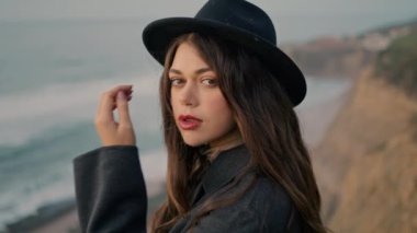Gentle young woman portrait standing in front gloomy seashore looking camera. Stylish gorgeous girl in black hat posing nature alone enjoying beautiful ocean view close up. Relax tranquility concept.