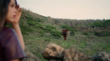 Unknown long-haired woman looking cow eating green grass at beautiful field evening time. Young calm girl standing on gloomy nature watching cattle animal grazing close up. Relax tranquility concept.