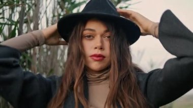 Portrait of fashion gorgeous model posing at high grass wearing black stylish hat. Close up attractive woman face with red lips long dark hair. Beautiful girl touching head standing near dry plants.