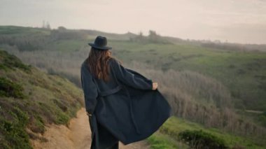 Back view young woman traveler walking rural road wearing cozy dark coat elegant hat. Long-hair attractive brunette going down dirty path looking back. Romantic girl tourist enjoy cloudy autumn day.