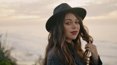 Relaxed girl tourist smiling touching long dark hair at cloudy sky backdrop close up. Attractive sensual lady traveler standing at coast overcast day. Happy playful woman model posing on nature alone.
