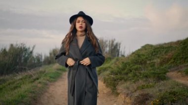 Serious attractive girl walking along dirt road unbuttoning dark coat. Confident long-haired woman in elegant hat going on rural path near green plants overcast autumn day. Young lady looking nature.