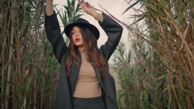 Stylish attractive girl standing high grass wearing black hat close up. Attractive young woman raising hands posing near dry plants cloudy day. Portrait of serious pretty brunette model looking camera