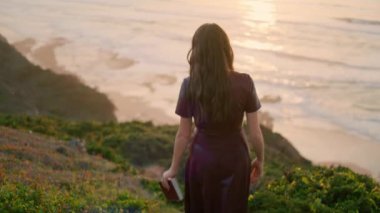 Back view dark-haired slim woman walking down green grass hill in front sunset seascape. Unknown young brunette holding book going to beach. Romantic girl relaxing on beautiful nature. Rest concept.
