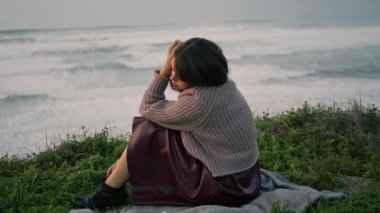 Sad lonely girl sitting on blanket with book enjoying beautiful sea view at dusk. Attractive young woman relaxing on seashore wearing gray sweater. Tranquil melancholic brunette feeling loneliness.