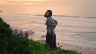 Pensive model posing at overcast seashore wearing cozy sweater. Attractive calm woman looking camera standing on green grass coast. Attractive romantic girl brunette relax enjoying evening nature.