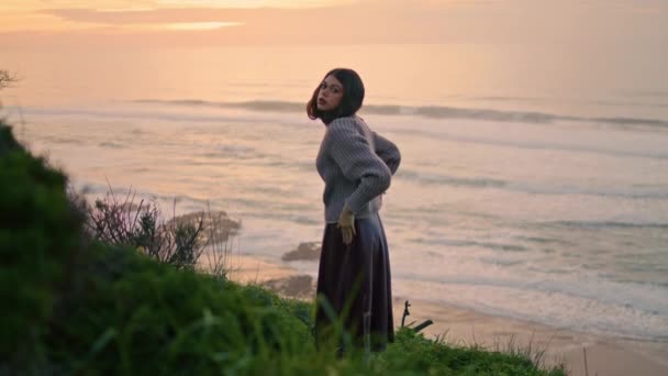 居心地の良いセーターを着て曇りの海岸でポーズを取るパッシブモデル 緑の芝生の海岸に立ってカメラを見て魅力的な穏やかな女性 魅力的なロマンチックな女の子ブルネットは夜の自然を楽しんでリラックス — ストック動画