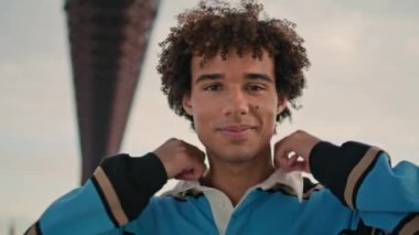 Smiling teenager posing camera at street fashion portrait. Positive zoomer standing looking alone closeup. Curly hair guy demonstrating emotions at city settings. Casual student face staring lens