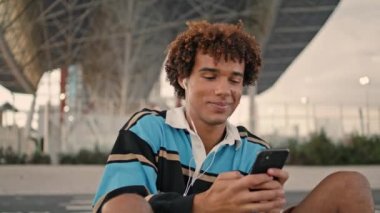 Curly student smiling smartphone at street closeup. Happy teenager browsing social media at mobile phone outdoors. Laughing earphones blogger enjoying music at evening. Joyful guy chatting friend 