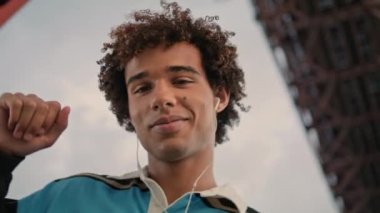 Earphones teen dancing street portrait. Young man listening music at cloudy sky background. Curly hair guy face looking camera at urban place. Active zoomer moving joyfully alone at city closeup 