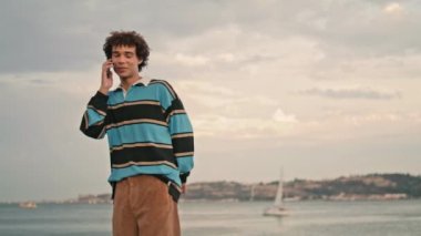 Smiling student walking mobile at river view place. Cheerful teen having pleasant cellphone call at windy day. Active hipster chatting smartphone. Happy young man touching hair. Phone talk concept 