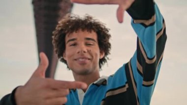 Positive student hands gesturing street portrait. Curly model photographing arms at urban bridge place. Smiling guy looking forward camera at cloudy sky background closeup. Happy teen face having fun